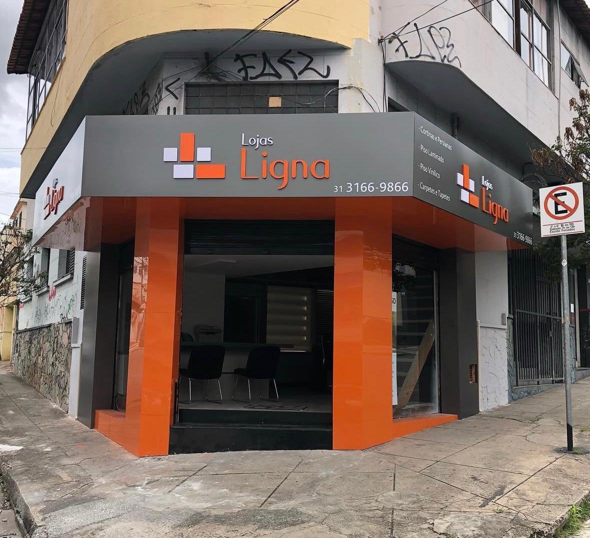 Fachada de esquina da Lojas Ligna com painéis cinza e colunas laranja em perspectiva urbana.